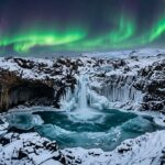 Frozen glacial waterfall and volcanic basalt cliffs under Northern Lights in winter Iceland