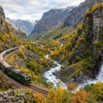 Panoramic view of the green vintage Flåm Railway train descending through the dramatic autumn valley of Flåmsdalen beside the roaring Kjosfossen waterfall in Norway