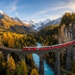 Panoramic Swiss red train crossing a stone viaduct in the Swiss Alps surrounded by autumn larch trees and snowy peaks