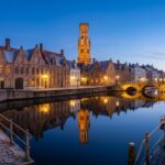Panoramic view of Rozenhoedkaai Quay of the Rosary in Bruges Belgium at winter twilight with historic Flemish brick guildhouses reflecting on calm canal water and the medieval Belfry tower