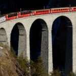 Panoramic view of the red Bernina Express train crossing the snowy Landwasser Viaduct high above alpine pines in Graubünden Switzerland