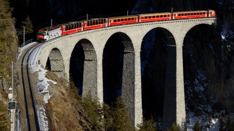 Panoramic view of the red Bernina Express train crossing the snowy Landwasser Viaduct high above alpine pines in Graubünden Switzerland