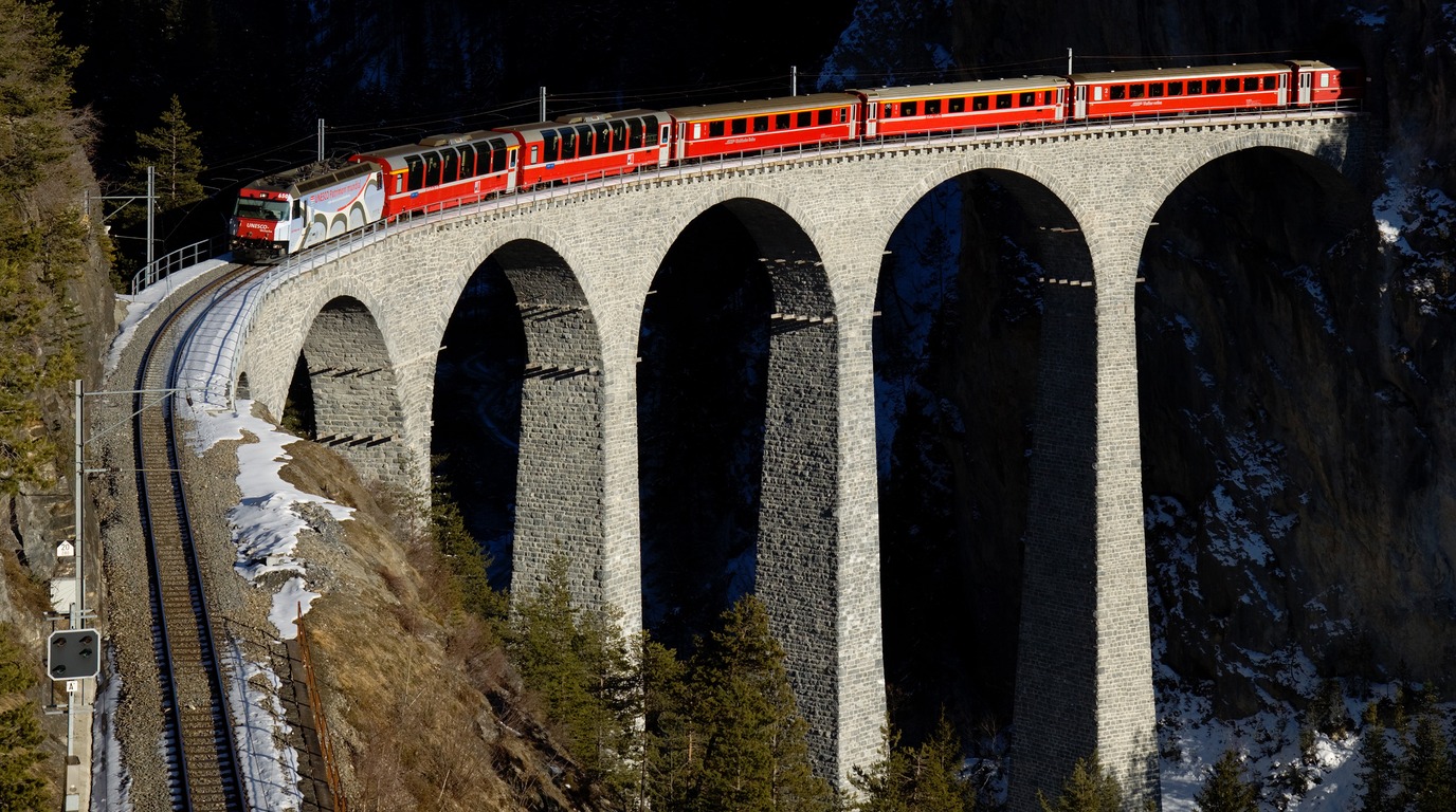 Red Bernina Express train crossing the dramatic snow-covered Landwasser Viaduct high above deep alpine pine valleys in Graubünden Switzerland