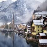 Panoramic winter view of Hallstatt Austria with historic 16th century Alpine wooden houses and church tower reflected in calm icy Lake Hallstatt beneath snow capped mountains