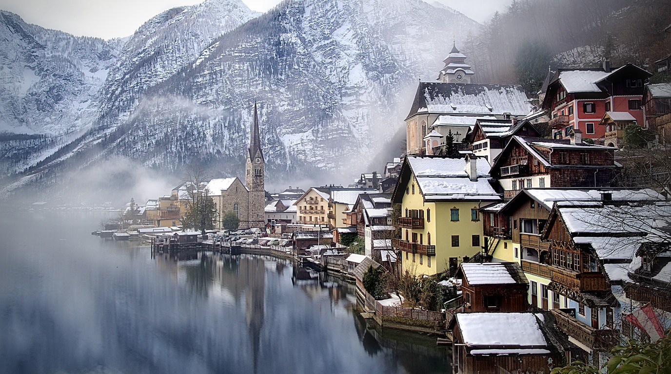 Panoramic winter view of Hallstatt Austria with historic 16th century Alpine wooden houses and church tower reflected in calm icy Lake Hallstatt beneath snow capped mountains