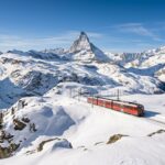 Red Gornergrat cogwheel train traveling through deep winter snowdrifts with the sharp peak of the Matterhorn mountain rising dramatically in Zermatt Switzerland